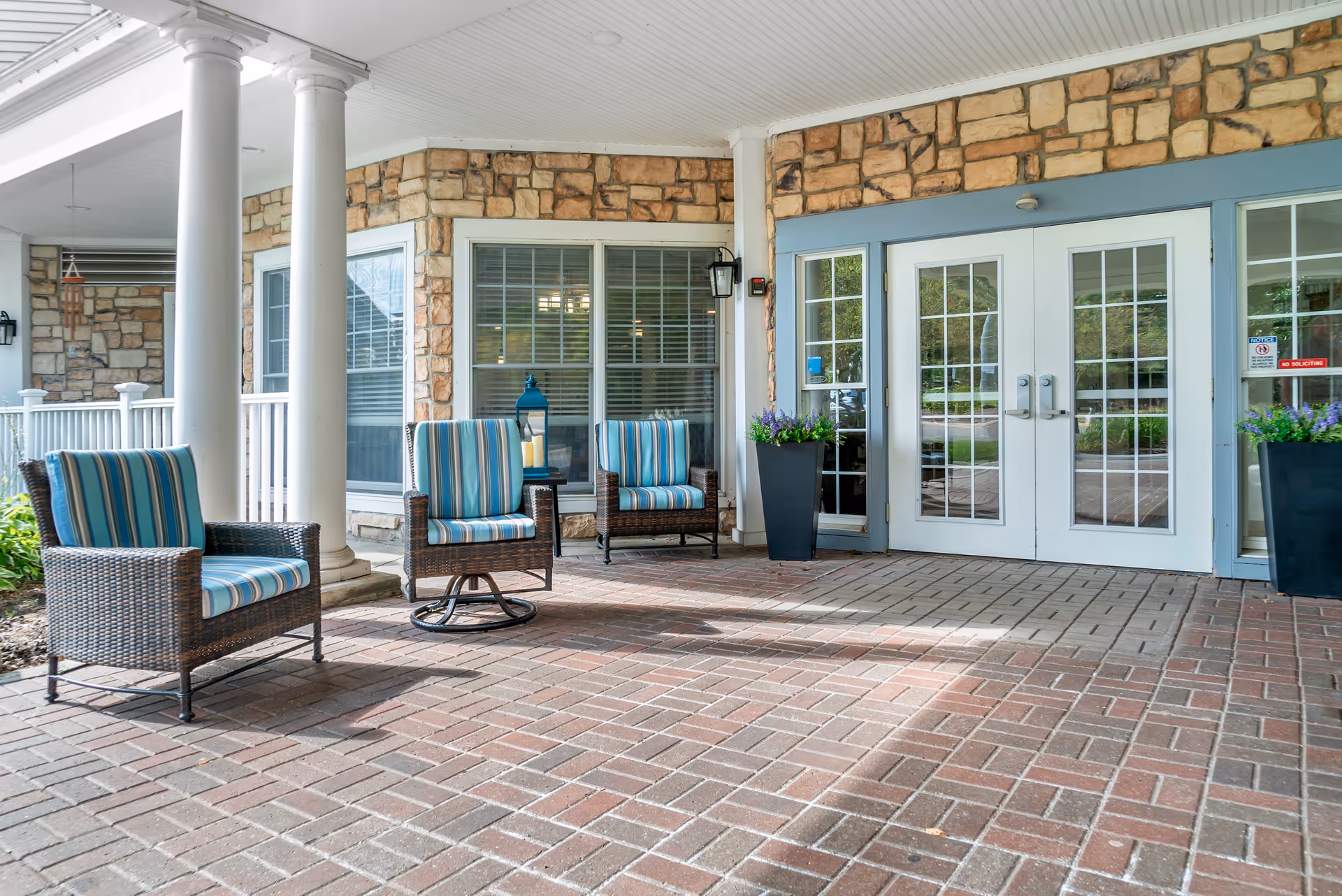Covered outdoor patio area with brick flooring, three wicker chairs with blue and white striped cushions, two tall black planters with purple flowers, stone walls, white columns, and double glass doors with white frames.