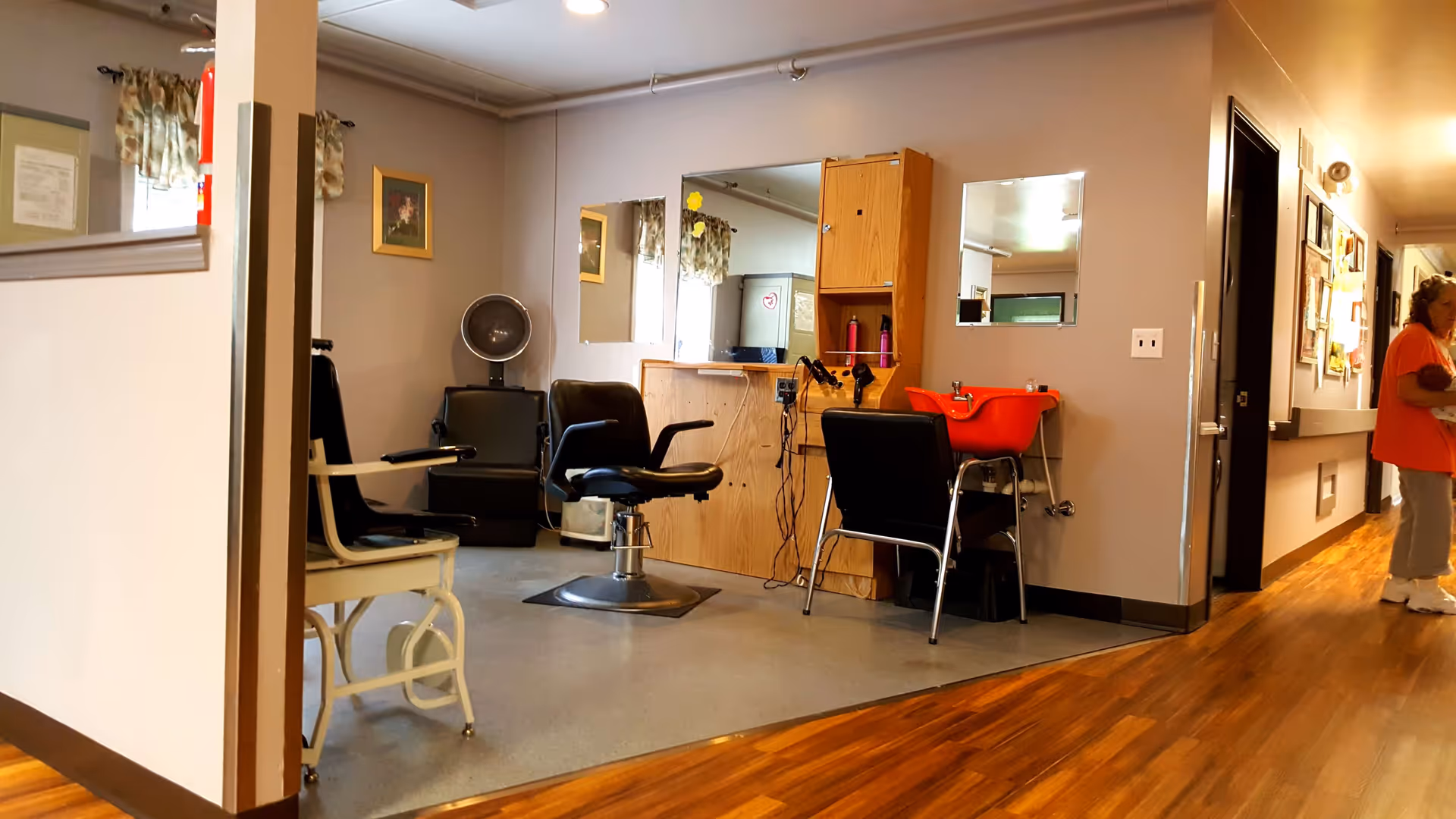 Interior view of a salon area in a senior living facility with a hair washing station, salon chairs, mirrors, and a hair dryer. A woman is standing in the hallway to the right, holding a baby doll. The floor is a mix of wood and linoleum, and the walls are decorated with framed pictures and bulletin boards.