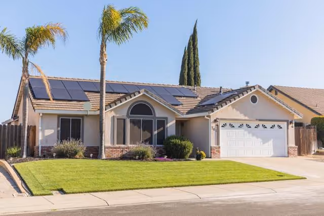 Single-story suburban house with a well-maintained green lawn, two tall palm trees, solar panels on the roof, and a two-car garage under a clear blue sky.