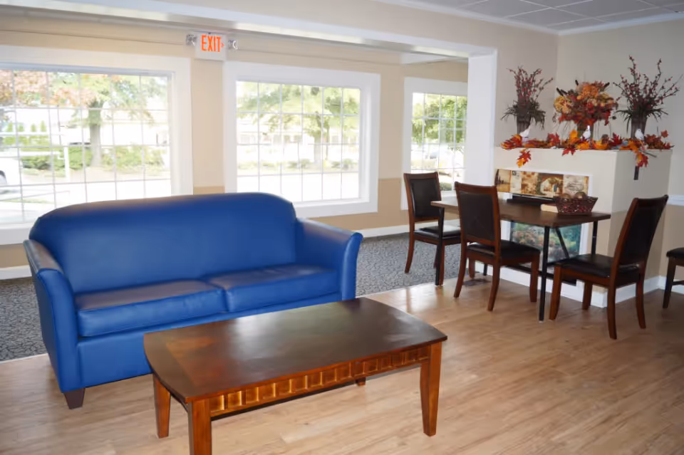 Bright common room with a blue sofa, wooden coffee table, and a small dining table with chairs by large windows.