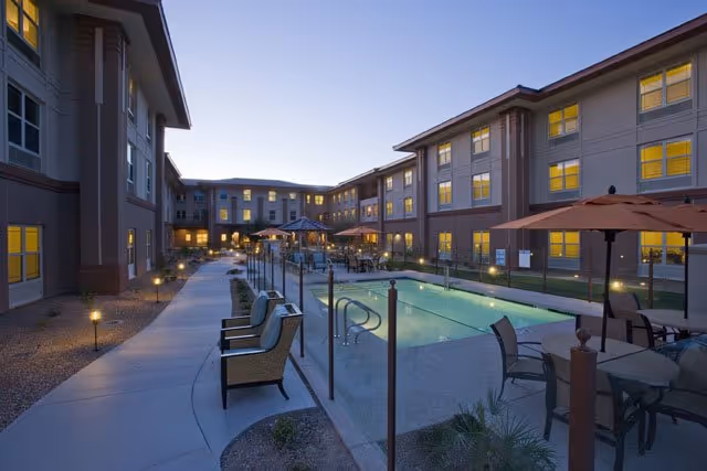 Outdoor courtyard area of a senior living facility at dusk featuring a swimming pool surrounded by a glass fence, patio tables with umbrellas, chairs, and a paved walkway with small garden beds and outdoor lighting. The multi-story building with many lit windows encloses the courtyard.