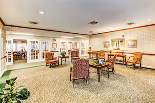 A spacious, well-lit common area with patterned carpet and wallpaper. The room contains several wooden chairs with cushions arranged around small tables. There are lamps on tables along the walls, framed artwork, and a clock above double glass doors leading to another room.