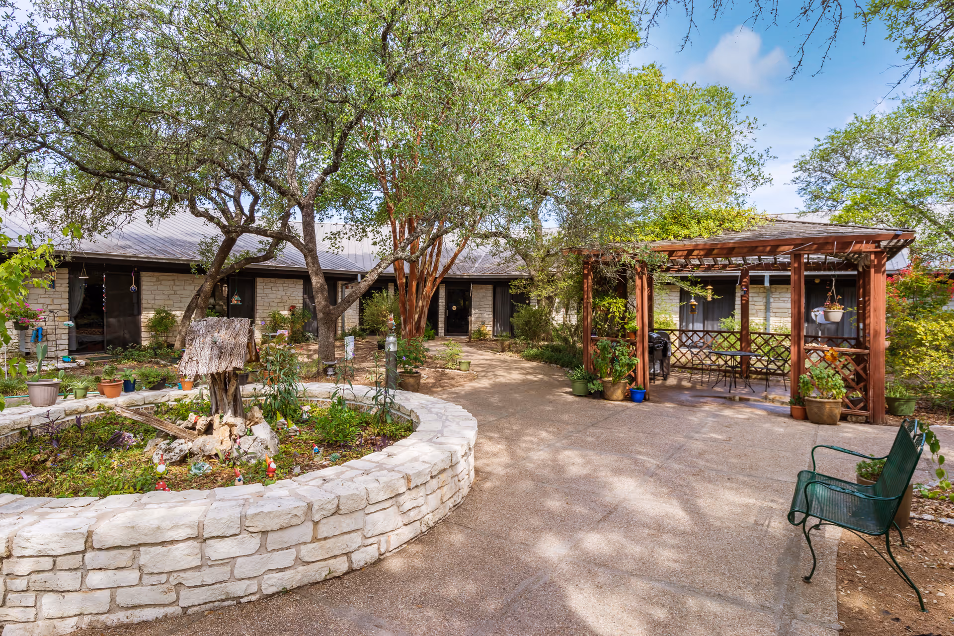 Outdoor courtyard area at Deer Creek Nursing and Rehab featuring a circular raised stone planter with various plants and small garden decorations. Surrounding the planter are mature trees providing shade. To the right, there is a wooden pergola with hanging plants and a small table with chairs underneath. A green metal bench is positioned on the paved walkway. The building exterior is visible in the background with stone walls and multiple doors and windows.