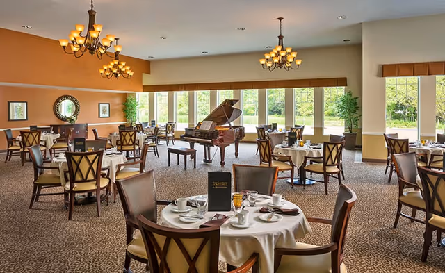 Spacious dining room with round tables set for meals, a grand piano in the center, chandeliers, and large windows overlooking greenery.