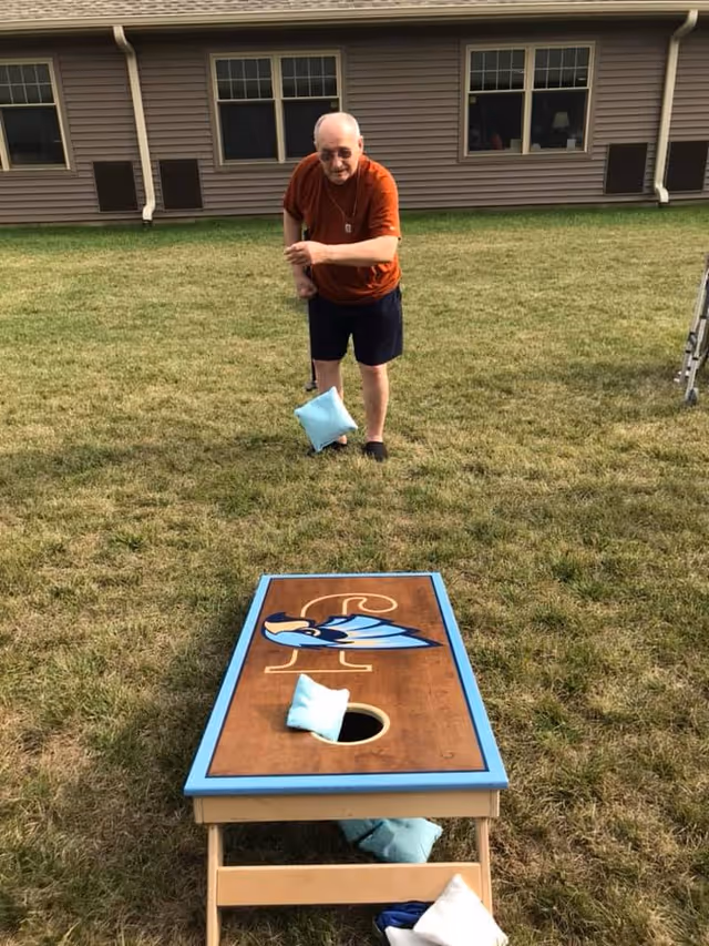 An elderly man wearing sunglasses, an orange shirt, and dark shorts is playing cornhole on a grassy lawn in front of a brown building with several windows. He is tossing a light blue bean bag towards a wooden cornhole board that has a blue bird design and a hole near the top. Several bean bags are on and around the board.