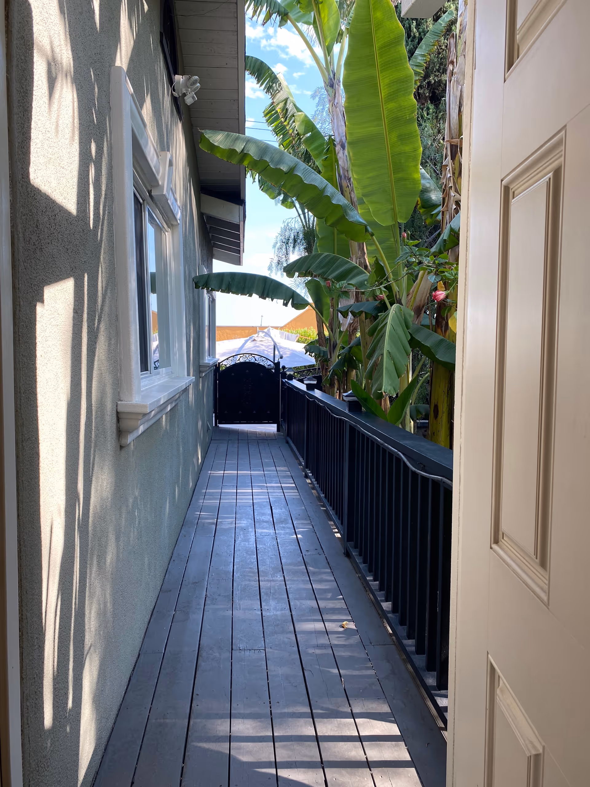 A narrow outdoor walkway with wooden flooring and a black railing on the right side. On the left side is the exterior wall of a building with windows. At the end of the walkway is a black gate, and large green tropical plants are visible on the right side beyond the railing. The sky is partly cloudy.