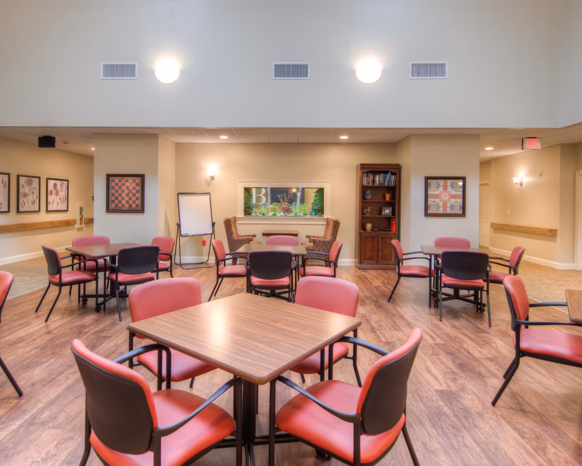 A well-lit common area with multiple square wooden tables surrounded by red and black chairs. The room has wood flooring, beige walls, framed artwork, a bookshelf, and two armchairs near a window with plants visible outside. There is also a whiteboard on a stand and ceiling lights providing illumination.