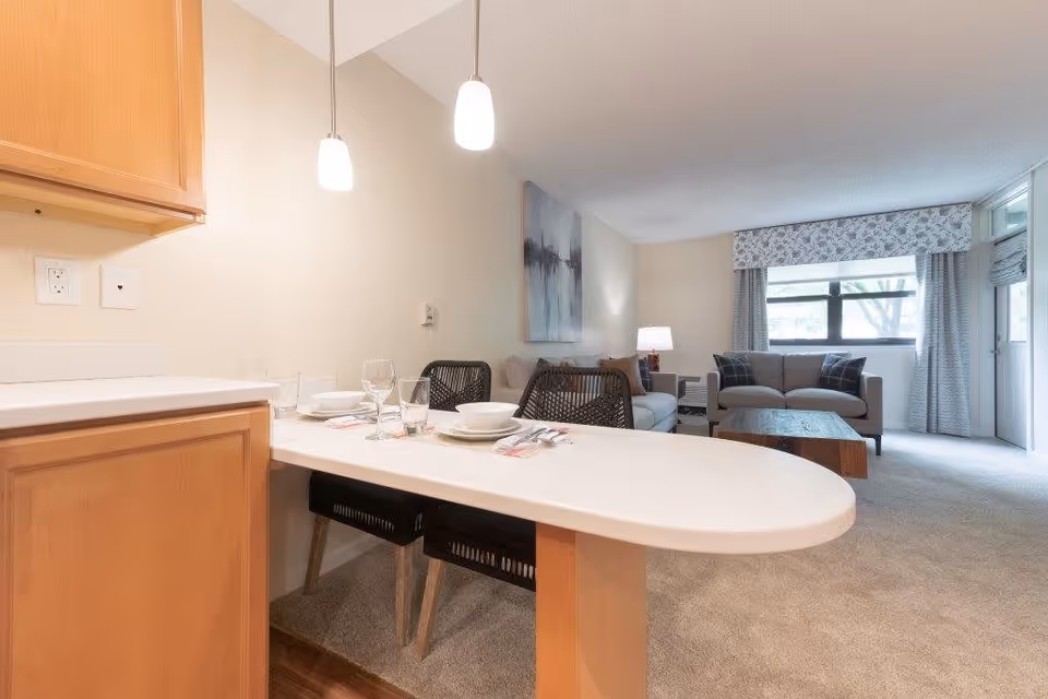 View of a cozy living room and dining area in an apartment at The Lafayette. The foreground shows a white countertop with place settings for two, including plates, glasses, and utensils. Two pendant lights hang above the counter. In the background, there are two sofas with cushions, a wooden coffee table, a side table with a lamp, and a large window with patterned curtains letting in natural light.