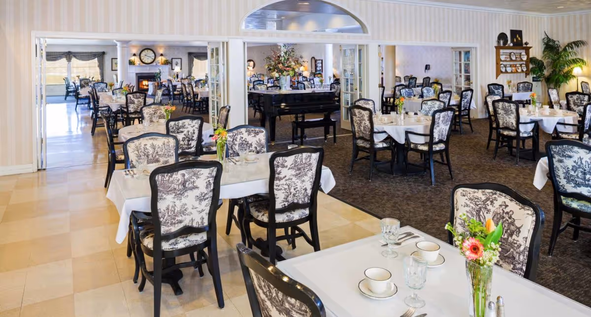 A spacious dining room in an assisted living community with multiple tables covered in white tablecloths, each set with cups, glasses, and silverware. The chairs have a black frame with patterned upholstery. There is a grand piano in the background, floral centerpieces on the tables, and a fireplace with a clock above it in an adjoining room.