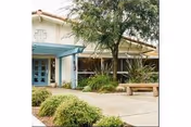 Exterior view of a senior living facility named Pacific Coast Manor showing a building entrance with large windows, a blue pergola, a tree, shrubs, and a wooden bench on a paved walkway.