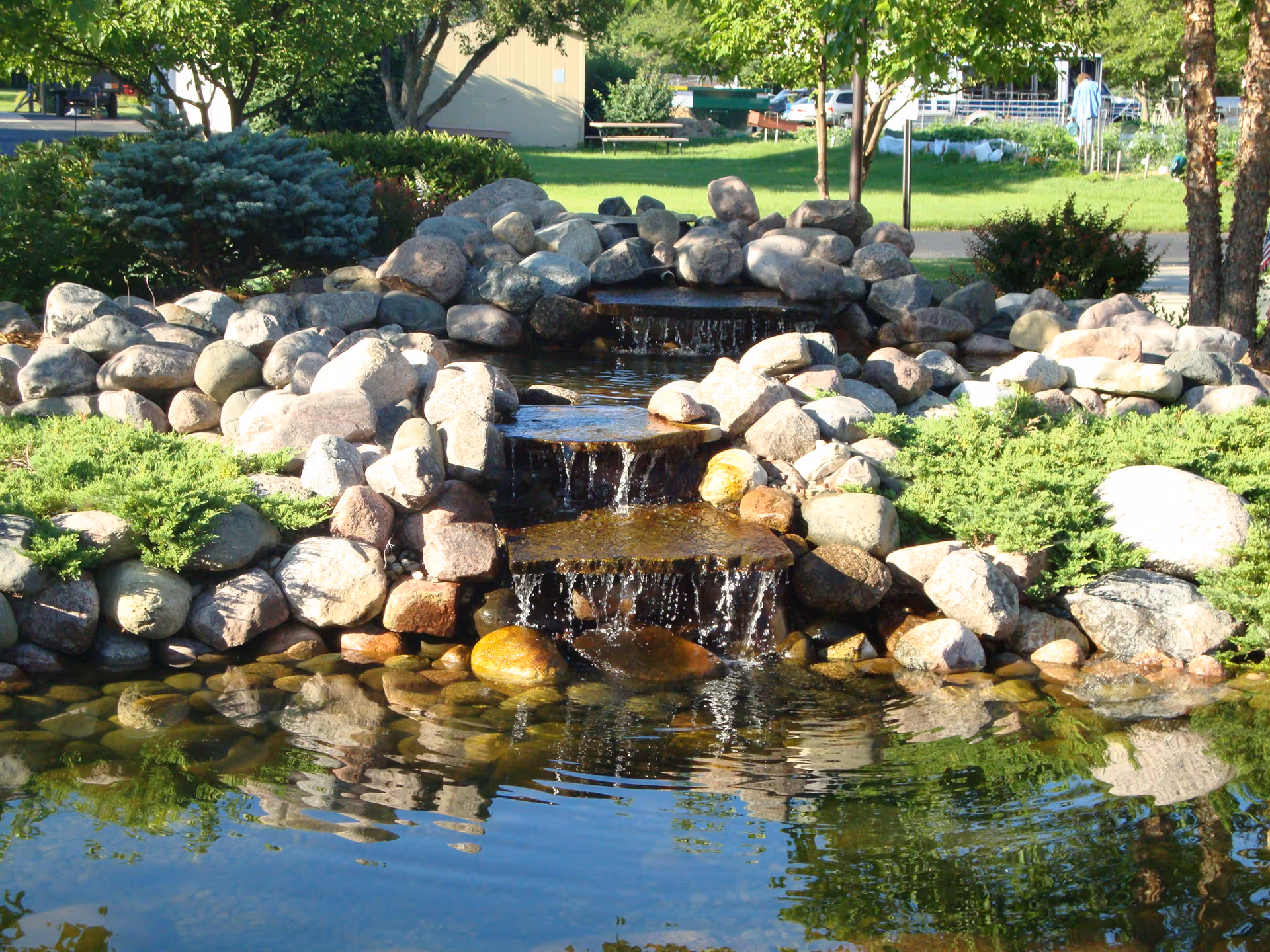 A small landscaped outdoor water feature with a cascading waterfall flowing over rocks into a pond, surrounded by green shrubs and trees in a garden setting.