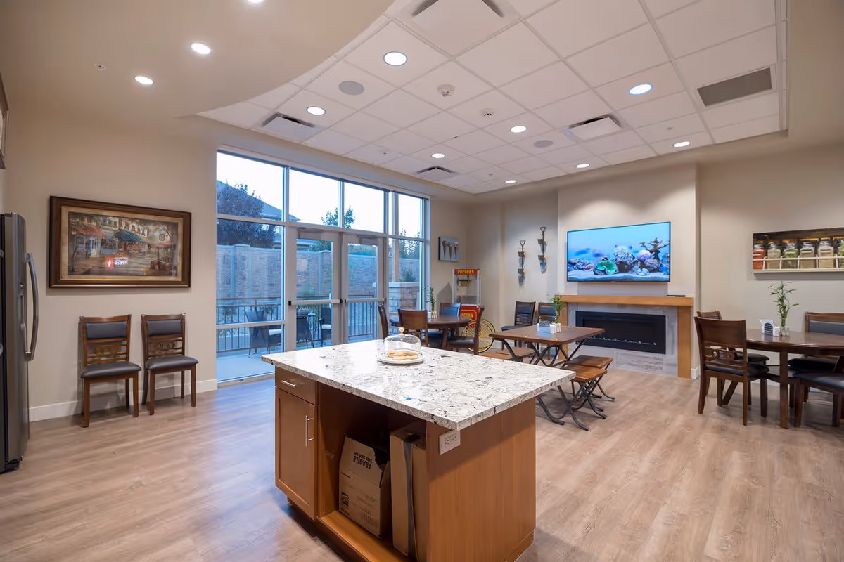 A bright and spacious common area in Crescent Senior Living featuring a kitchen island with a granite countertop, wooden chairs, and tables arranged for dining or socializing. Large windows and glass doors provide natural light and a view of an outdoor patio. A flat-screen TV is mounted above a modern fireplace, and a popcorn machine is visible in the corner. The room has light wood flooring and recessed ceiling lights.