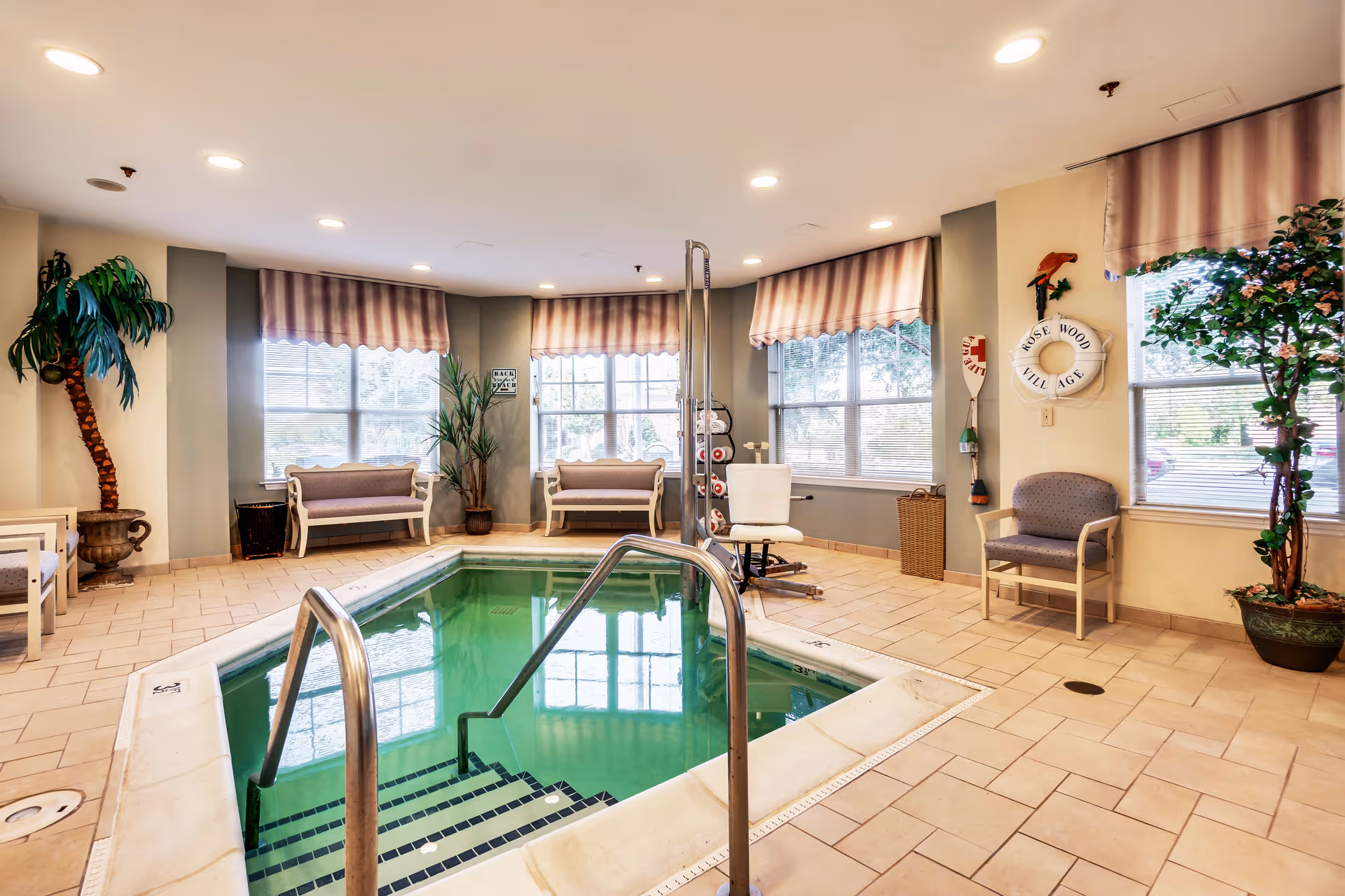 Indoor pool area with green water, metal handrails, and steps leading into the pool. The room has large windows with striped valances, several chairs and benches along the walls, potted plants, and a pool lift for accessibility. A life preserver on the wall reads 'Rose Wood Village'.