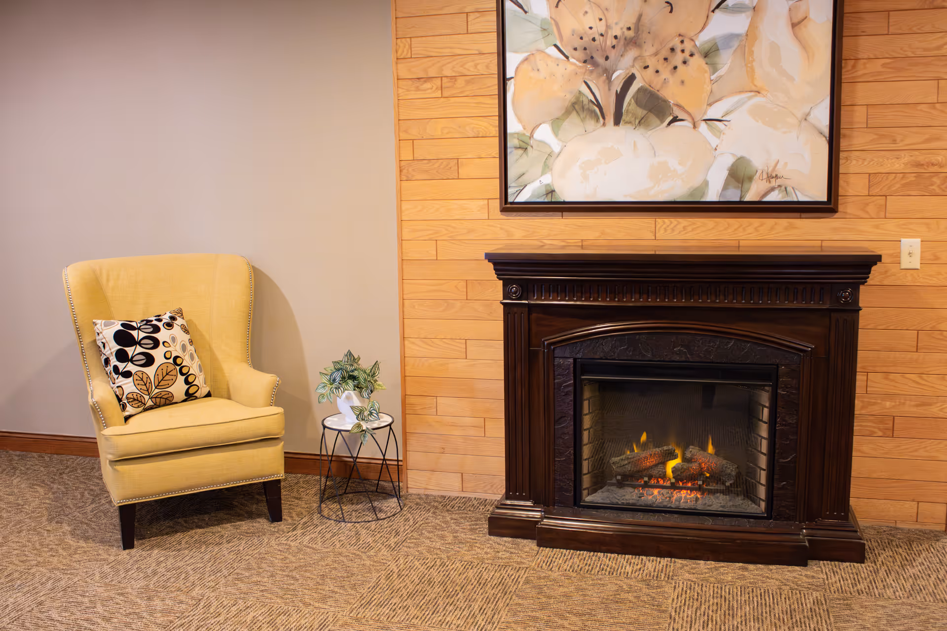 A cozy corner of a senior living facility featuring a yellow armchair, a decorative pillow, a small plant, and a fireplace.