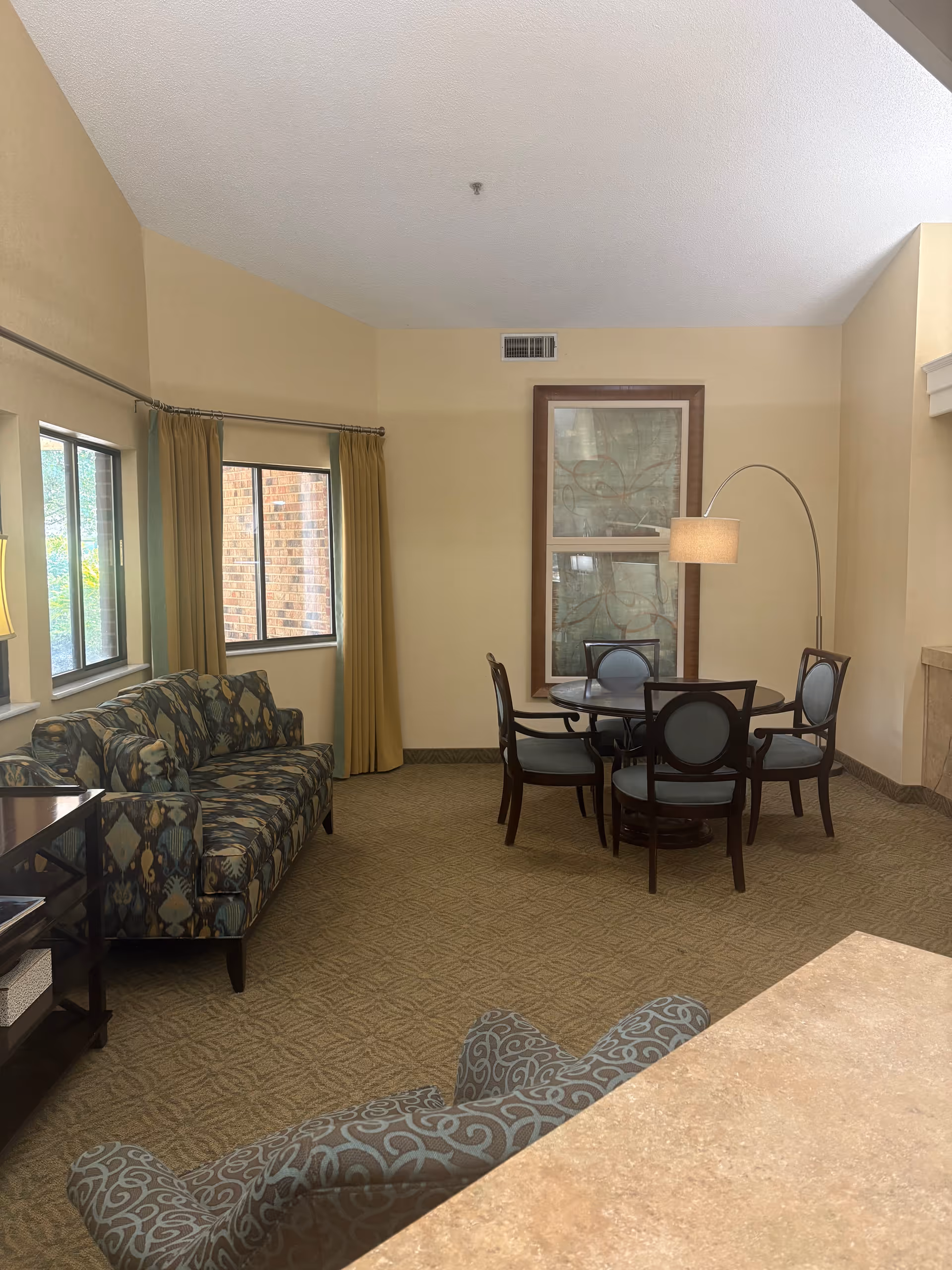 Comfortable senior living lounge with a patterned sofa and armchair, a round dining table with chairs, and a floor lamp in a carpeted room.