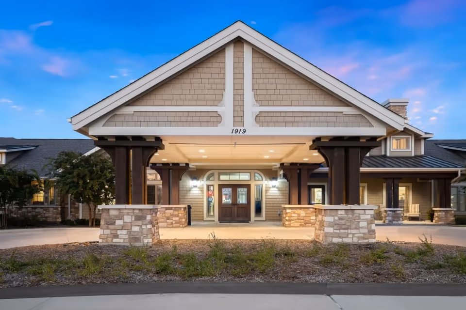 Front entrance of a senior living building with a covered porte-cochère, stone pillars, double doors, and a peaked roof against a twilight sky.