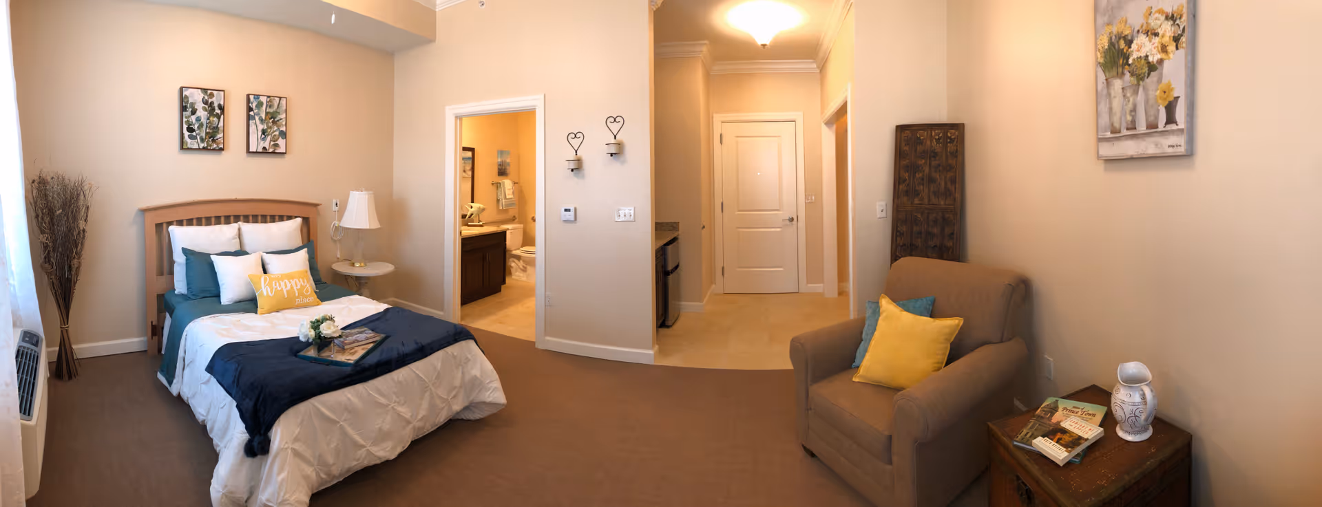A cozy senior living bedroom with a neatly made bed featuring white and teal bedding and a yellow pillow that says 'happy place'. There is a small round side table with a lamp next to the bed, a comfortable armchair with yellow and teal pillows, and a wooden chest with books and a decorative vase. The room has beige walls with framed botanical artwork and a floral painting. An open door reveals a bathroom with a vanity, toilet, and towel rack. The entrance hallway and a small kitchenette area with a mini fridge are also visible.