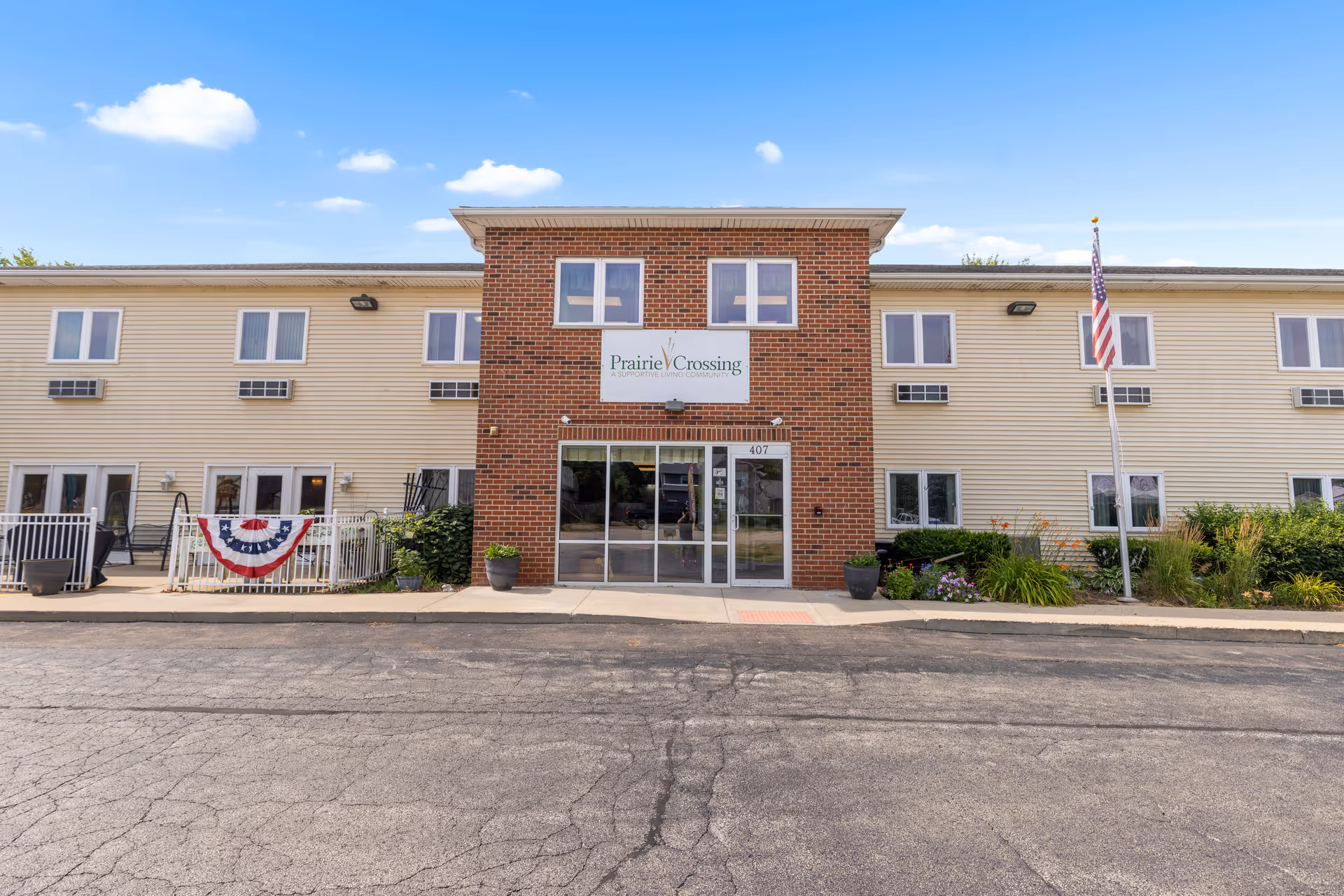 Front entrance of the Prairie Crossing Assisted Living building with a central brick entry, glass doors, an American flag, and landscaping.