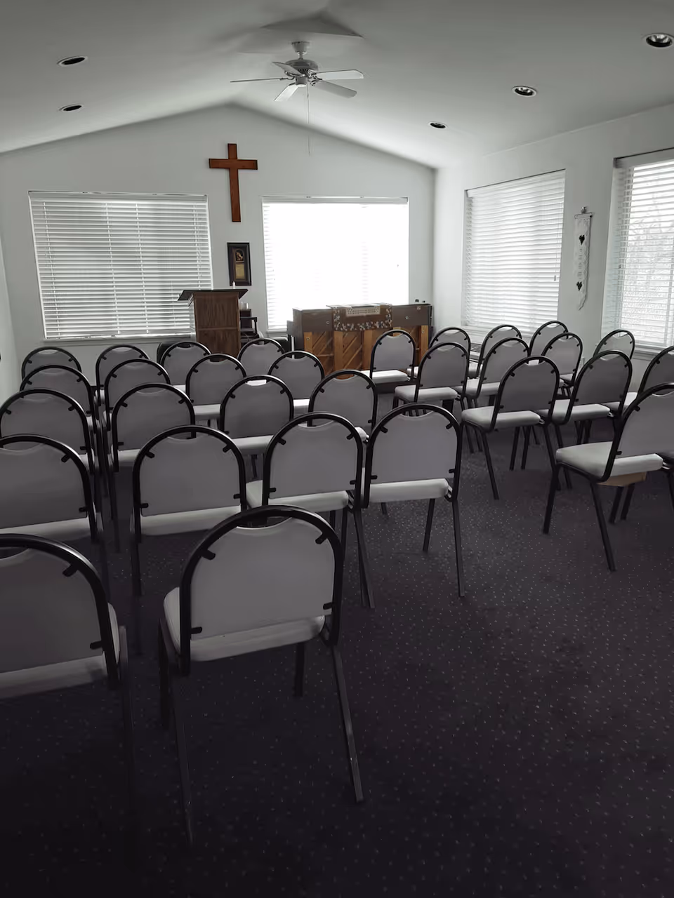 Interior view of a small chapel or meeting room with rows of chairs facing a wooden podium and a piano. A large wooden cross is mounted on the wall behind the podium. The room has white walls, three large windows with blinds, and a ceiling fan.