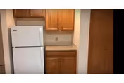 Small kitchenette with a white refrigerator, wooden upper and lower cabinets, a countertop, and a closed wooden door.
