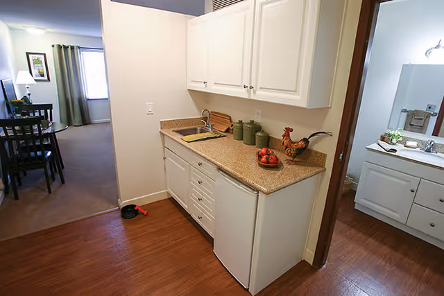 Small kitchenette with white cabinets, a countertop and sink opening to a dining area and a visible bathroom vanity.