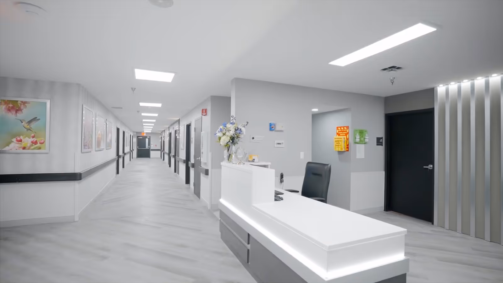 Well-lit rehabilitation facility interior showing a white reception desk in the foreground and a long corridor with doors and artwork along both sides.