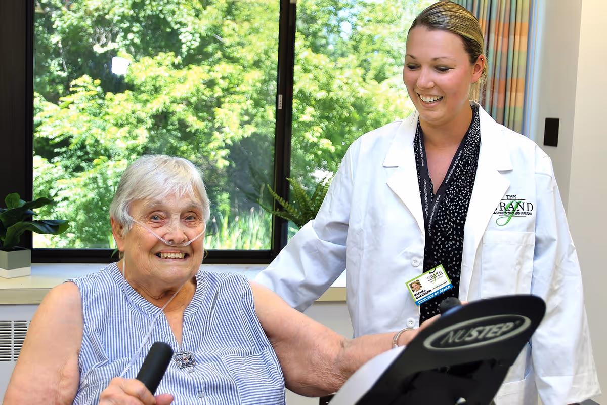 An elderly woman with a nasal oxygen tube is using a NuStep exercise machine indoors near a large window with green trees outside. A smiling female healthcare professional in a white coat with The Grand Rehabilitation and Nursing logo stands beside her, offering support.