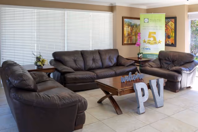 A bright lounge with brown leather sofas arranged around a wooden coffee table displaying a 'welcome PV' sign and a Palomar Vista banner.