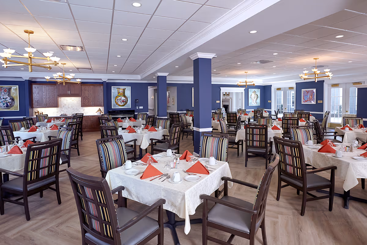 A spacious dining room with multiple tables covered in white tablecloths, each set with white cups, plates, silverware, and red folded napkins. The chairs have dark wood frames with striped upholstery. The walls are painted blue and decorated with framed artwork. The room has a light wood floor and modern gold chandeliers hanging from a white ceiling.