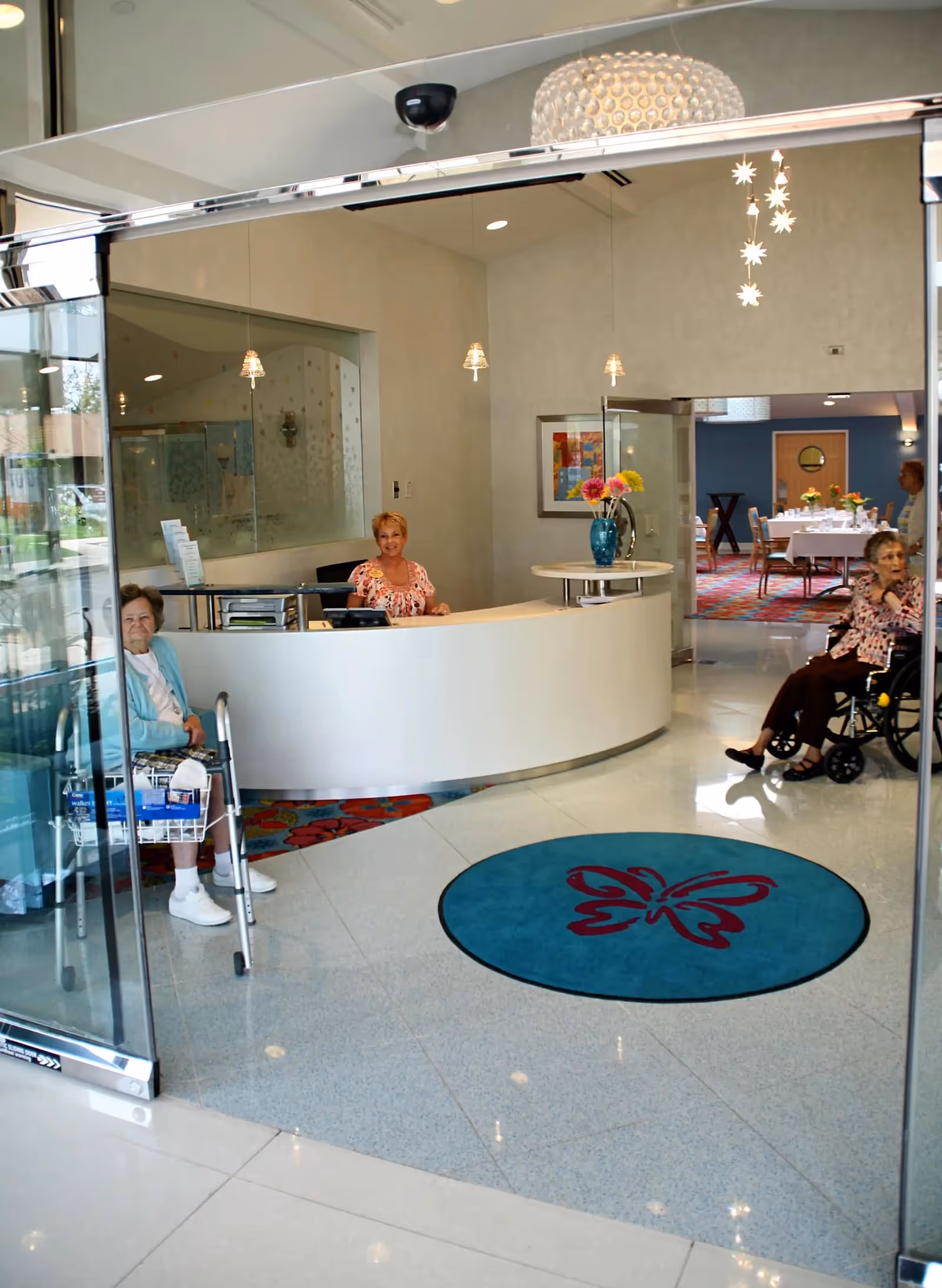 View of a senior living facility reception area with a curved white front desk where a receptionist is seated. Two elderly women are visible, one sitting with a walker near the entrance and another in a wheelchair. The floor has a blue circular rug with a butterfly design. In the background, there is a dining area with tables and chairs, and a vase with flowers on a small table near the reception desk.