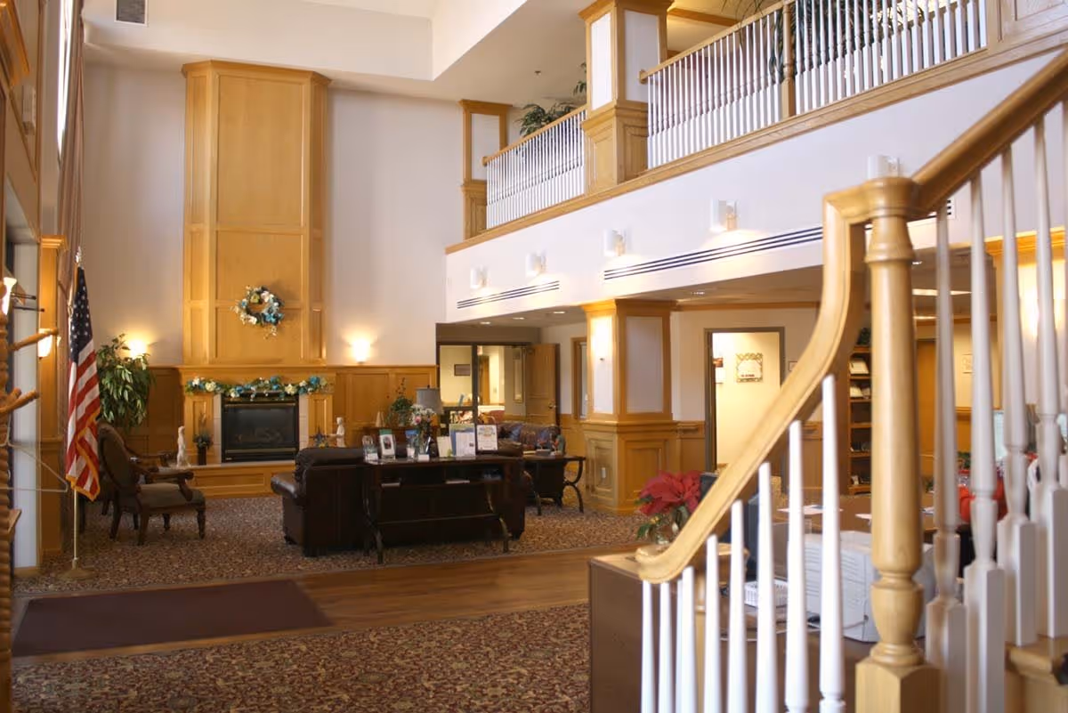 A spacious and well-lit common area with a high ceiling and wooden paneling. The room features a fireplace decorated with a wreath and garland, comfortable seating including leather sofas and armchairs, an American flag, and a staircase with wooden handrails and white balusters leading to an upper level balcony.