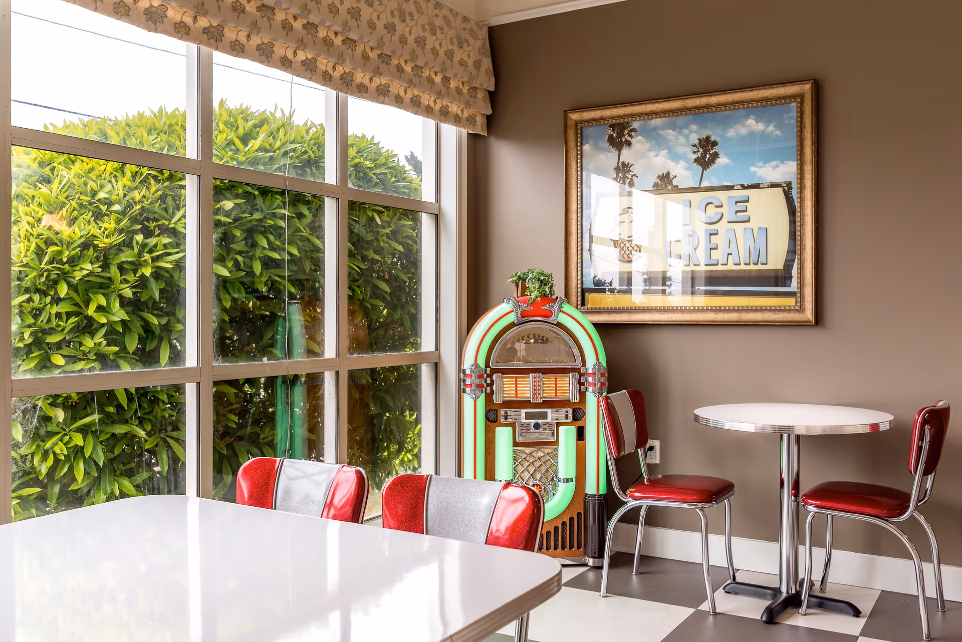 A bright dining area with a large window showing green bushes outside. Inside, there is a retro-style jukebox with neon green lights, a framed picture of an ice cream sign on the wall, and two sets of tables and chairs with red and silver upholstery.