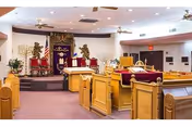 Interior view of a synagogue or chapel with wooden pews, a raised platform with a Torah ark covered by a purple curtain adorned with gold lions and Hebrew text, menorahs on either side, and ceiling fans above.