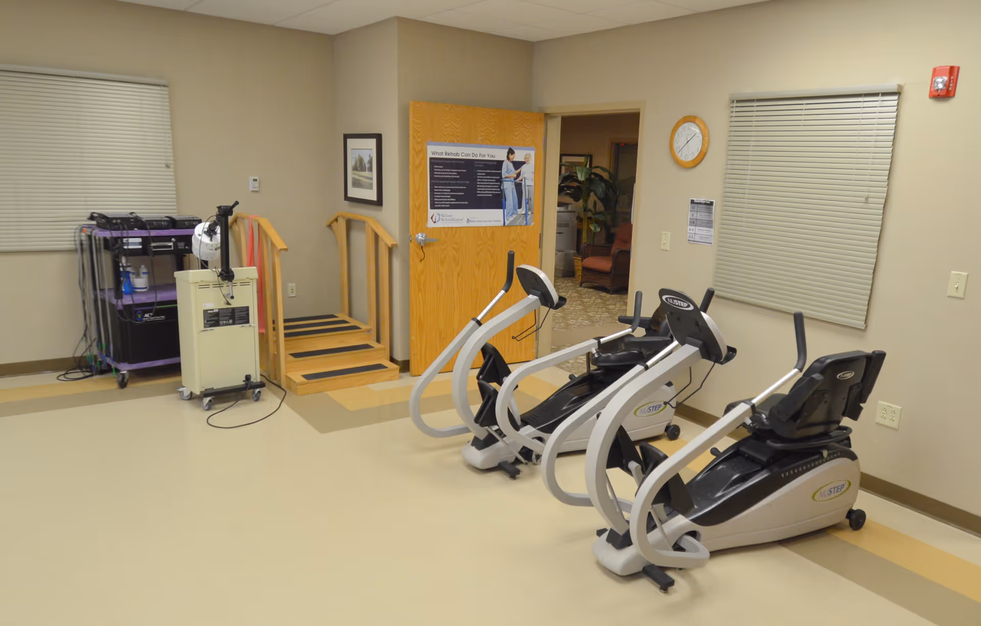 A rehabilitation exercise room with two NuStep recumbent cross trainers, a small set of wooden stairs with handrails, a medical equipment cart, and a door leading to another room. The walls are beige with two windows covered by blinds, a clock, and a poster on the door.