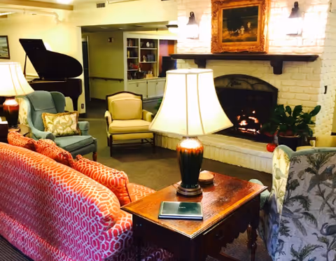 A cozy living room area with a lit fireplace set in a white brick wall. The room features a red patterned sofa, a blue armchair, a beige armchair, and a floral armchair arranged around a wooden side table with a lamp and a closed book. A grand piano is visible in the background along with built-in shelves and soft lighting.