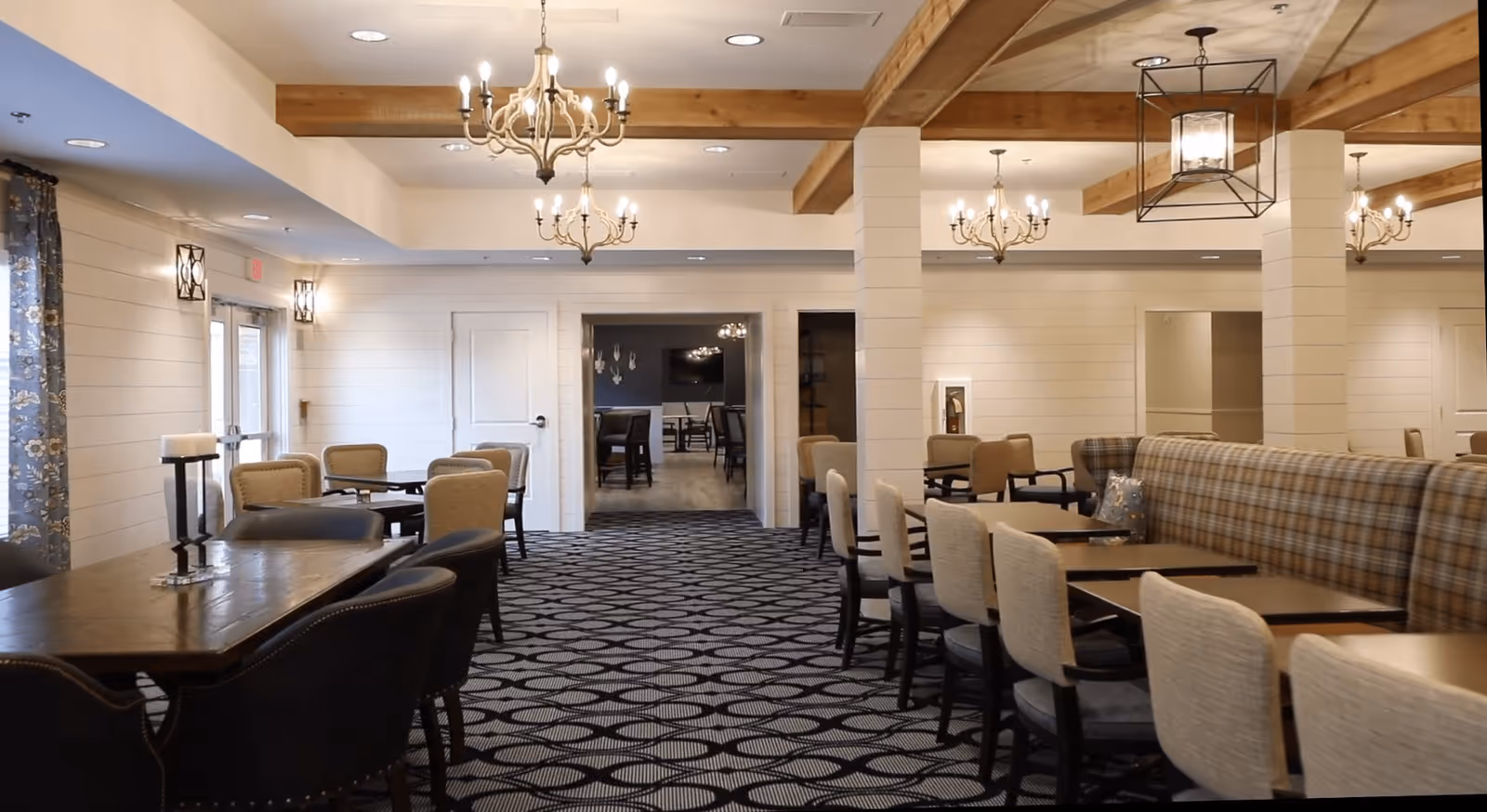 A spacious dining room with multiple tables and chairs arranged neatly. The room features patterned carpet flooring, wooden beams on the ceiling, and several chandeliers providing warm lighting. There are large windows with floral curtains on the left side, and a doorway leading to another room in the center background.
