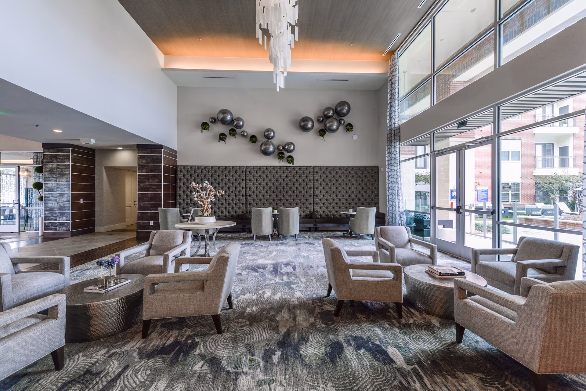A spacious and modern living room area in a senior living facility with multiple gray upholstered armchairs arranged around two oval coffee tables. The room features a high ceiling with a contemporary chandelier, large windows letting in natural light, and a wall decorated with metallic spherical art pieces and small plants. There are additional seating areas with small tables and chairs along the back wall.