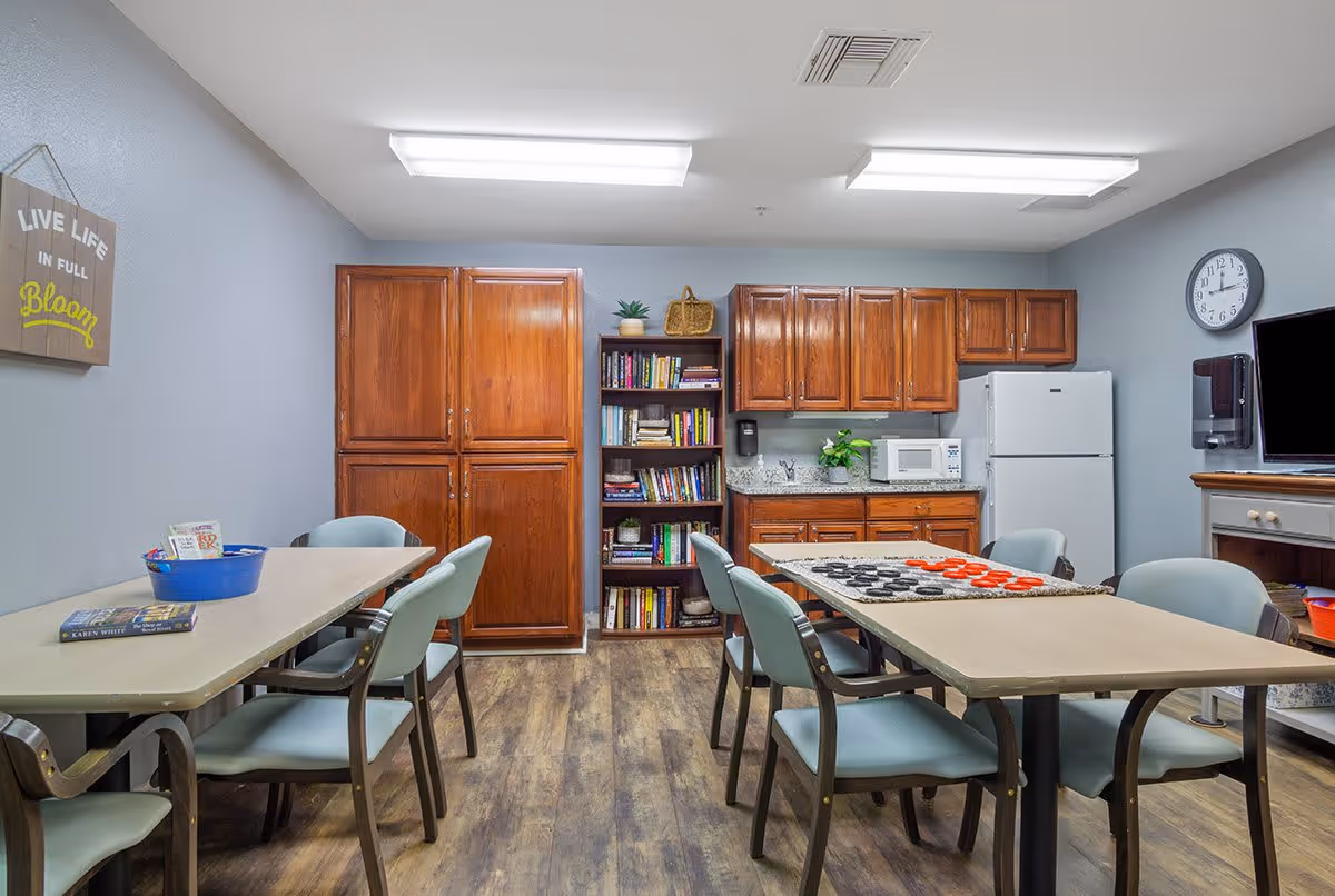 A cozy common room with two tables and several chairs arranged around them. One table has a large checkers board set up, and the other has a blue basket and a book on it. The room has wooden cabinets, a bookshelf filled with books, a microwave, a refrigerator, a wall clock, and a TV mounted on a stand. A sign on the wall reads 'LIVE LIFE IN FULL Bloom'. The floor is wooden, and the walls are painted light gray.