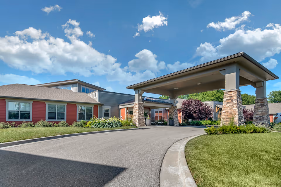 Exterior view of Carnegie Village Rehabilitation & Health Care Center showing a covered entrance supported by stone pillars, a driveway, well-maintained landscaping with green grass and shrubs, and a partly cloudy blue sky.