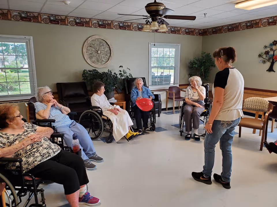A group of elderly women, some in wheelchairs, seated in a circle in a common room with a caregiver standing nearby. The room has windows, plants, wall decorations, and a ceiling fan with lights.
