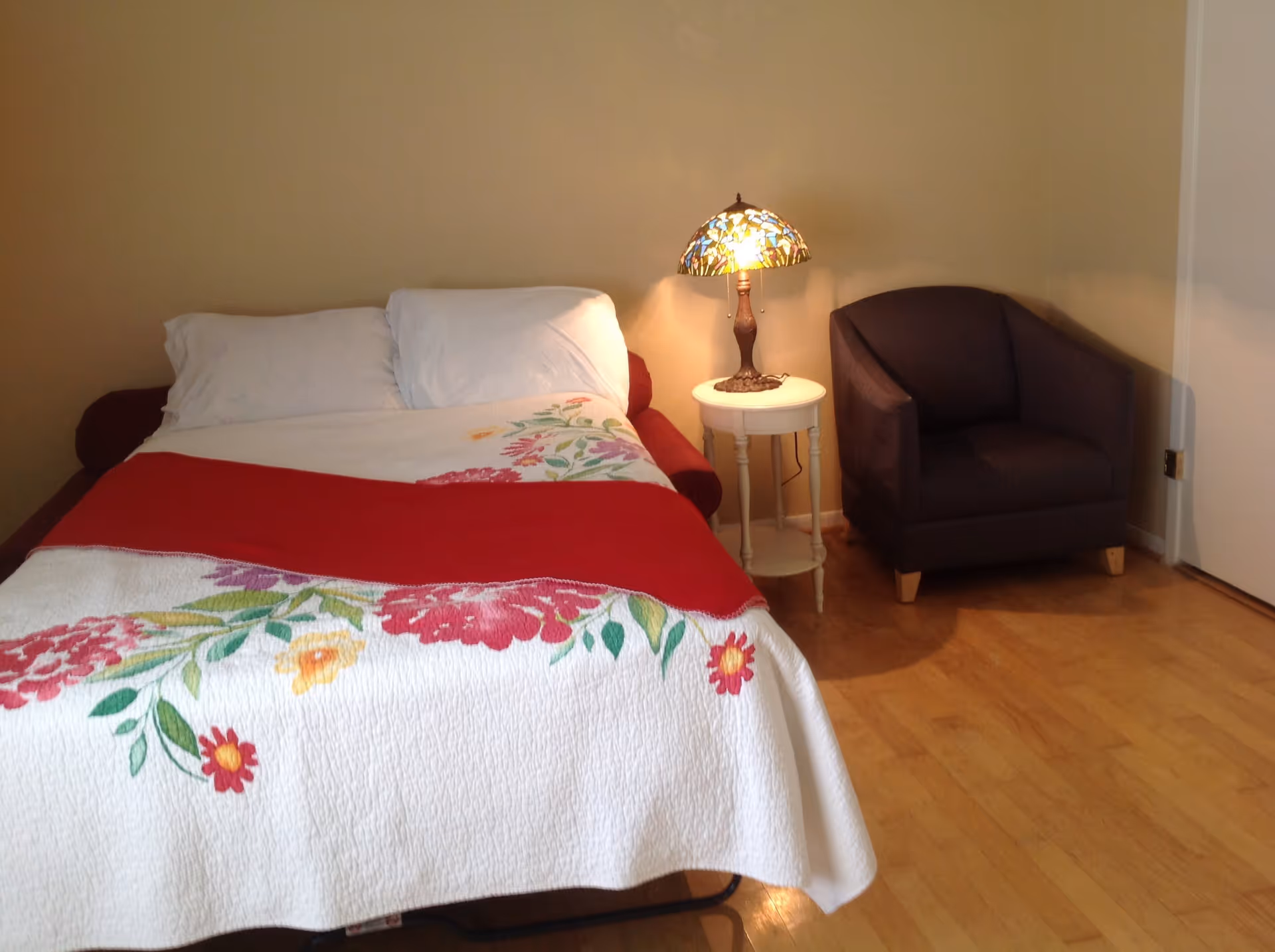 A cozy bedroom with a bed covered in a white quilt with red and floral patterns, two white pillows, a small round white side table with a decorative stained glass lamp, and a dark upholstered armchair on a wooden floor.