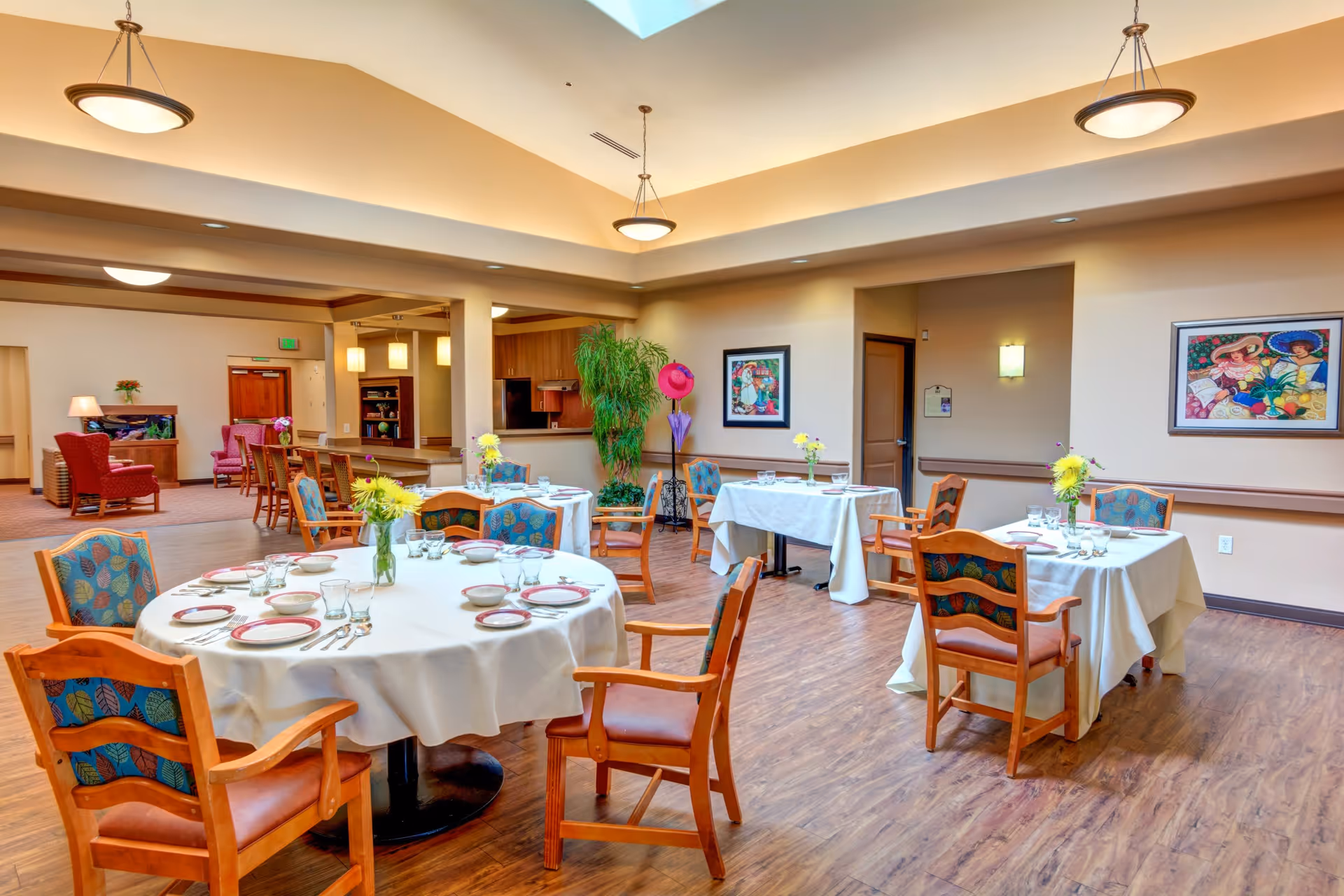 A bright and spacious dining room with round and square tables covered in white tablecloths, set with plates, bowls, glasses, and silverware. Each table has a small vase with yellow flowers. The room has wooden floors, colorful cushioned chairs, framed artwork on the walls, and warm overhead lighting. In the background, there is a seating area with red armchairs and a wooden cabinet.