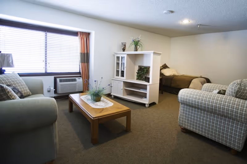 A cozy living area in a senior living facility with a green sofa and a plaid armchair surrounding a wooden coffee table with a plant centerpiece. A white entertainment cabinet separates the living space from a bed in the background. The room has a window with blinds and curtains, and a wall-mounted air conditioning unit below the window.
