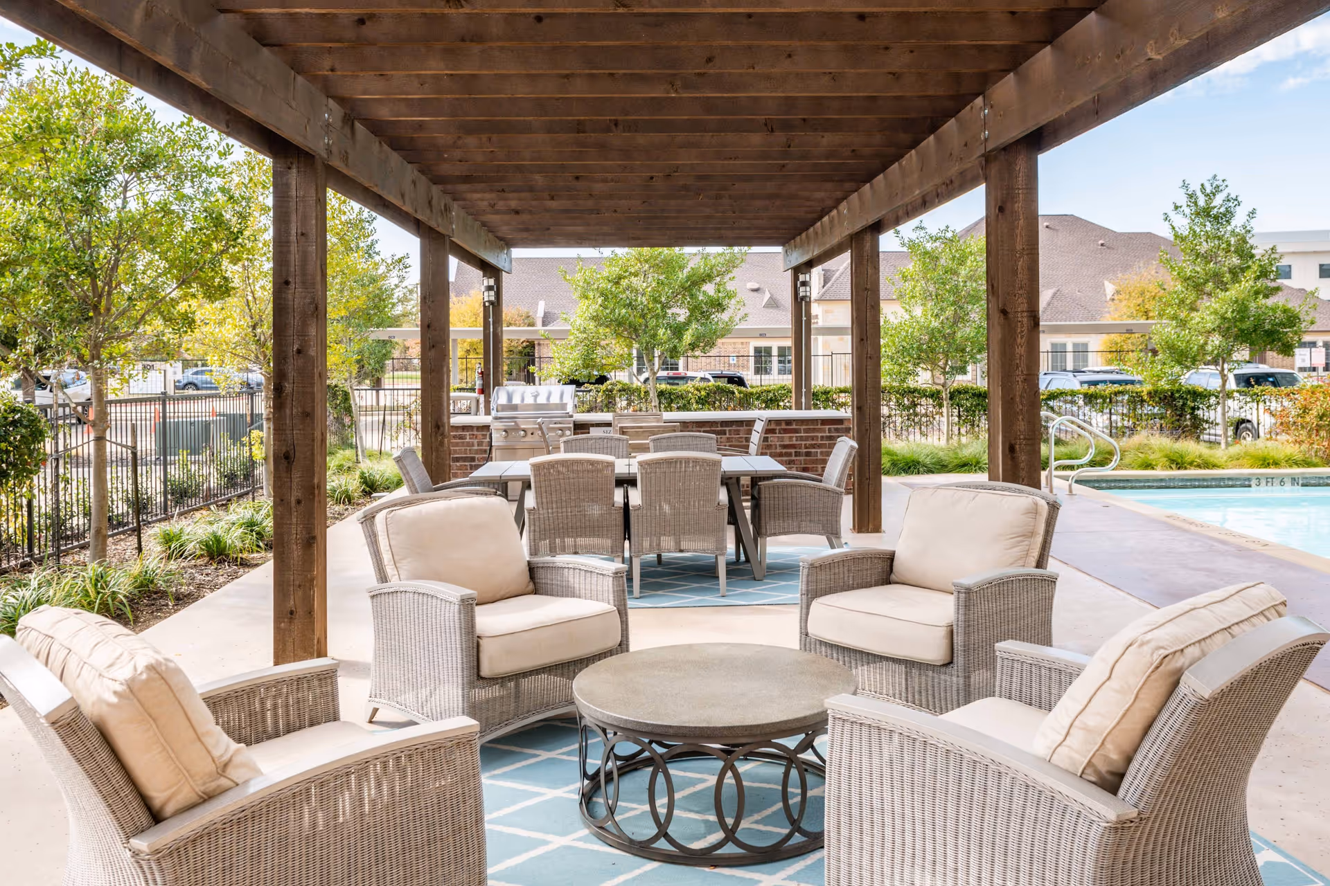 Outdoor covered seating area with four cushioned wicker armchairs arranged around a round metal and stone coffee table on a blue patterned rug. Behind the seating area is a dining table with six wicker chairs and a built-in grill. The area is shaded by a wooden pergola. In the background, there are trees, a fence, parked cars, and buildings under a partly cloudy sky.