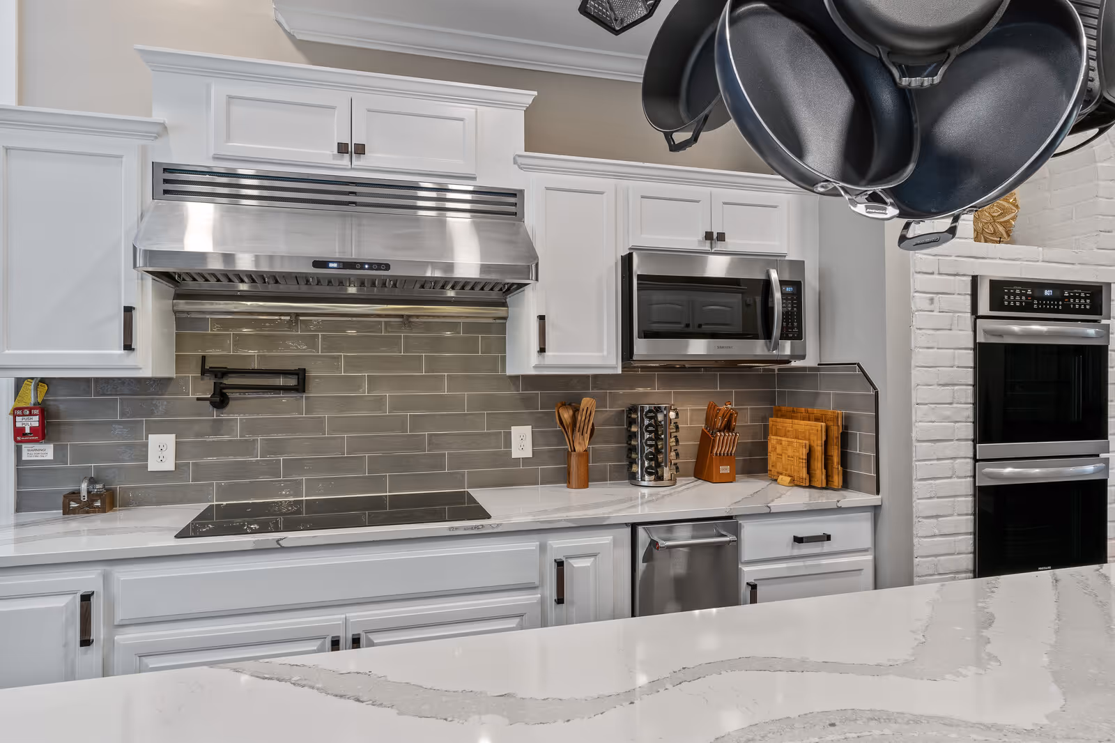 Modern kitchen with white cabinets, stainless steel appliances including a microwave, double oven, and stovetop with a stainless steel range hood. Gray subway tile backsplash and white marble countertops. Hanging pots and pans are visible in the upper right corner.