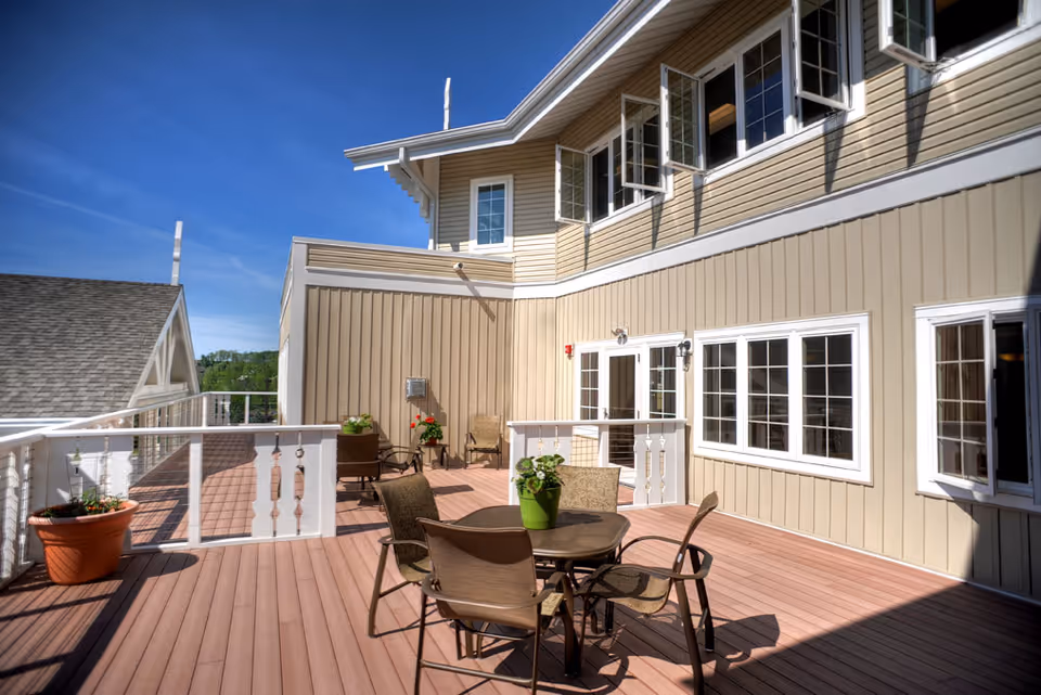 Sunlit outdoor deck with a round table and chairs, potted plants, and the beige exterior of a multi-story assisted-living building.