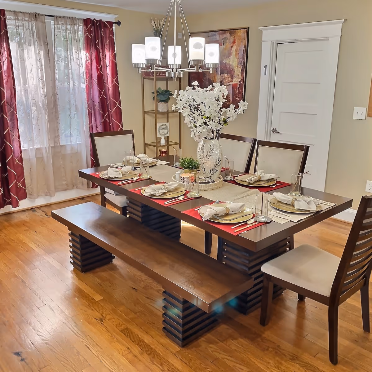 Dining room with a set wooden table, bench and chairs, chandelier lighting, and a floral centerpiece.