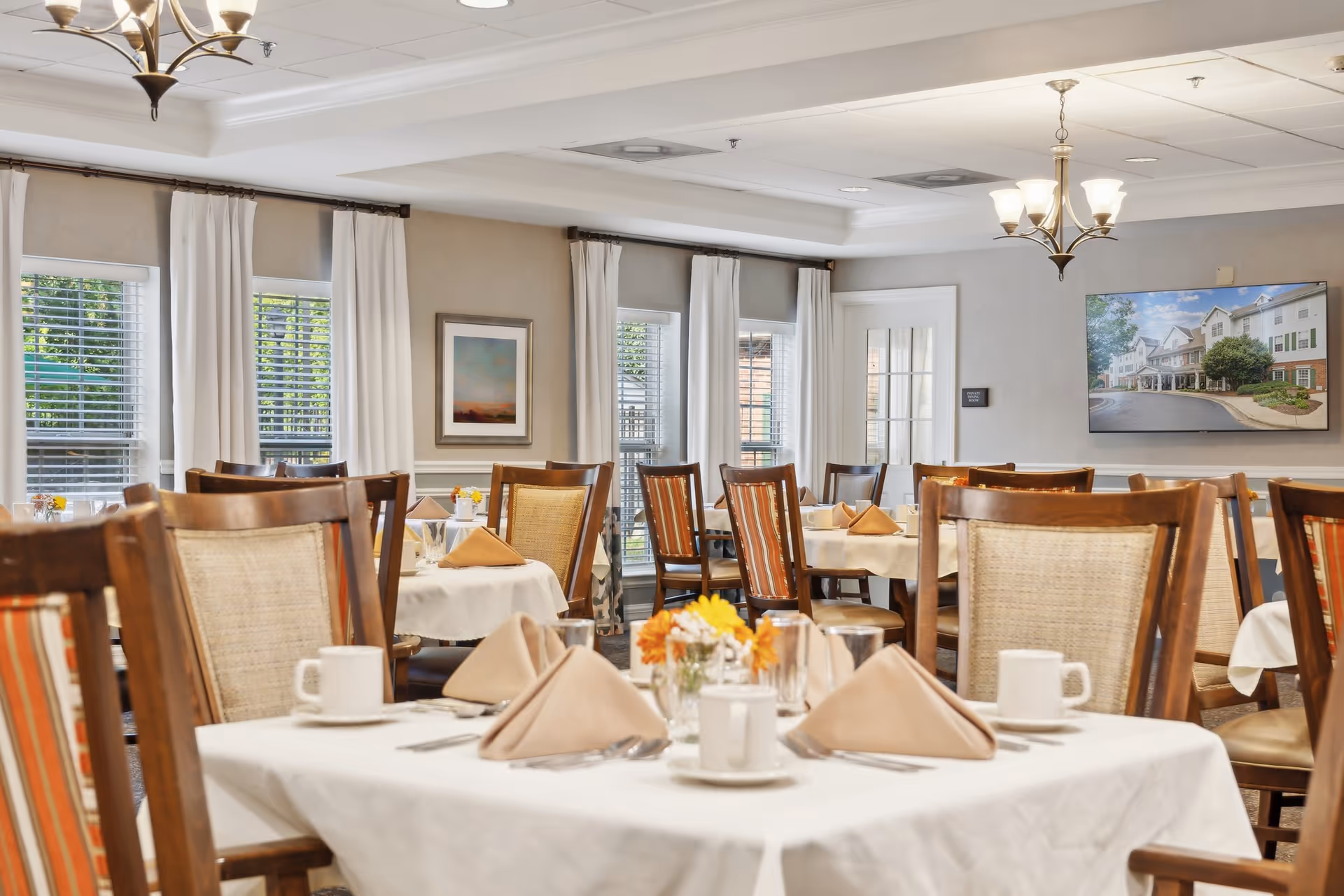 A bright and neatly arranged dining room with multiple tables covered in white tablecloths, each set with beige folded napkins, white coffee cups, glasses, and silverware. The room features wooden chairs with woven backs and striped cushions, large windows with white curtains, a chandelier, and framed artwork on the walls.