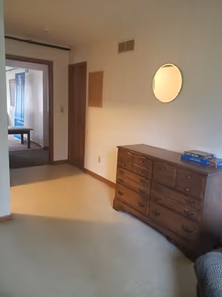 Interior view of a room with beige carpet and white walls. A wooden dresser with multiple drawers is positioned against the right wall beneath a round mirror. A doorway on the left leads to another room with a bench and a blue banner visible in the background.