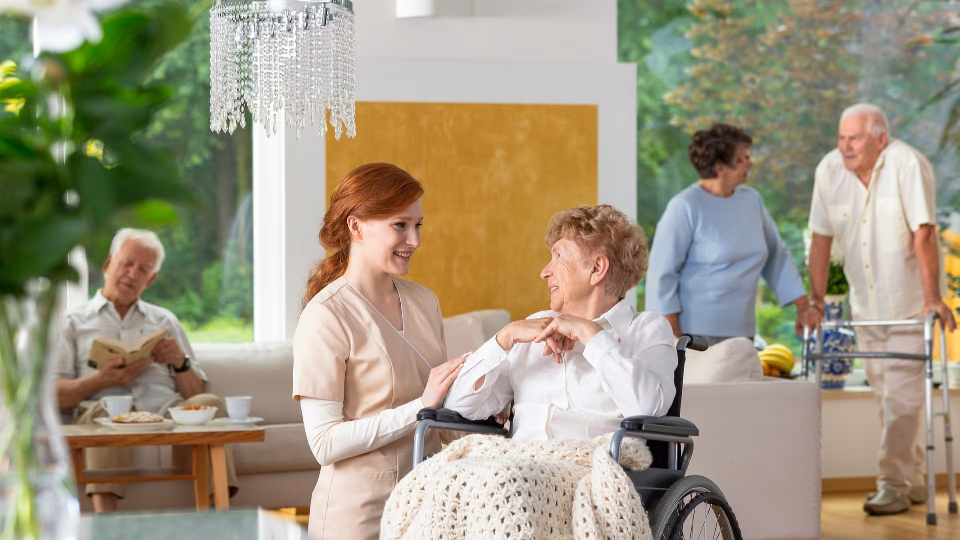 A caregiver smiles while talking with an elderly woman in a wheelchair in a bright communal living room with other seniors in the background.